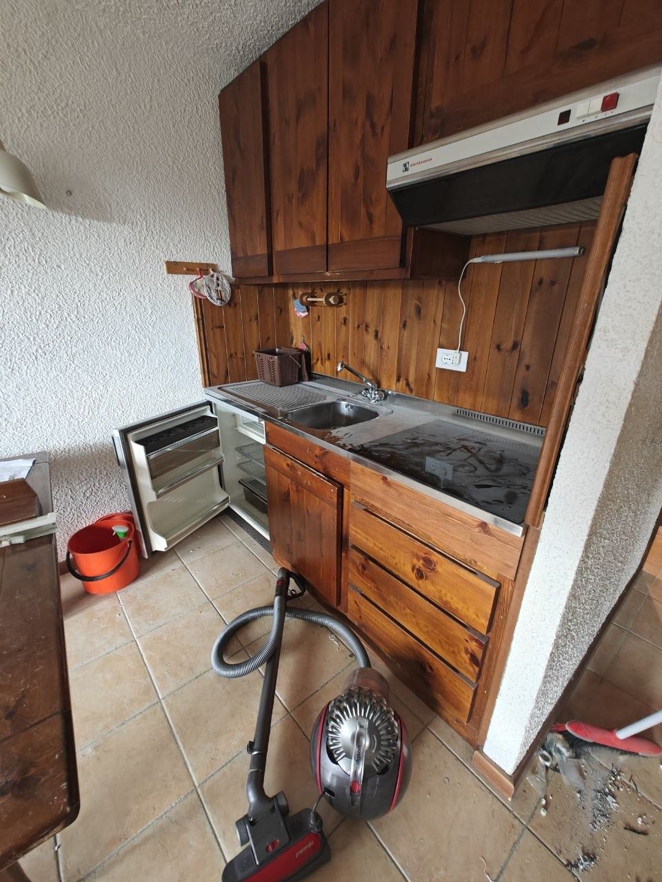 Angolo cottura in legno con frigorifero aperto, piano cottura sporco e detriti sul pavimento